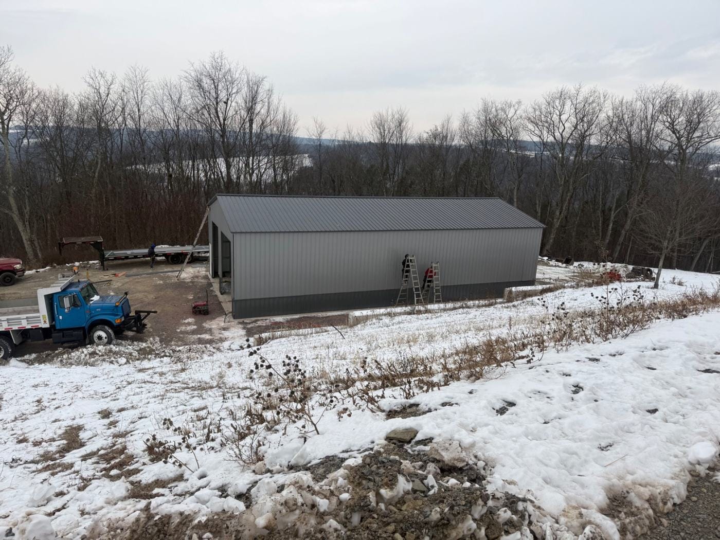 A metal building on a hilltop site in the snow, with a dump truck and plow at left.